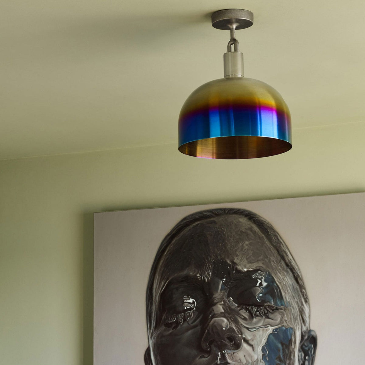 A ceiling light with a rainbow-colored shade hangs above a painting of a person's face covered in black liquid.