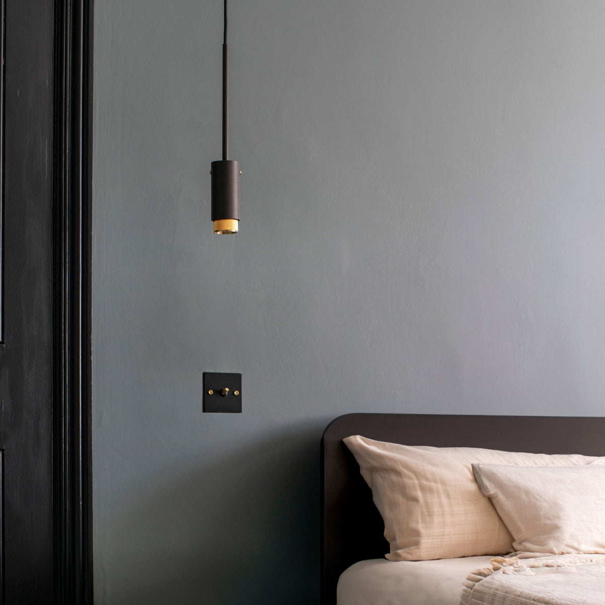 A modern bedroom with a dark grey wall, a black pendant light, and a bed with light-colored pillows.