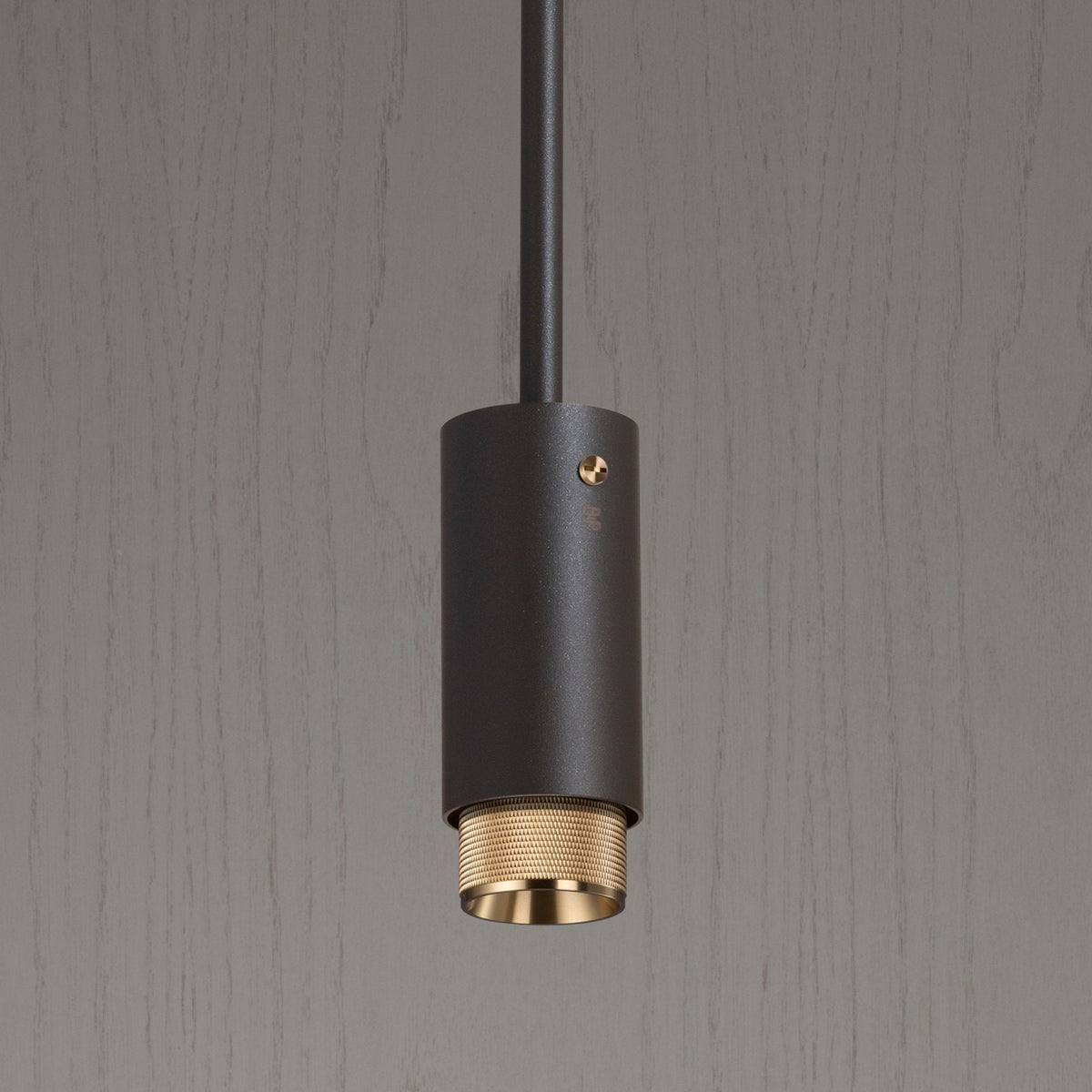 Close-up of a dark grey cylindrical pendant light with a textured brass base, against a light grey wood-grain background.