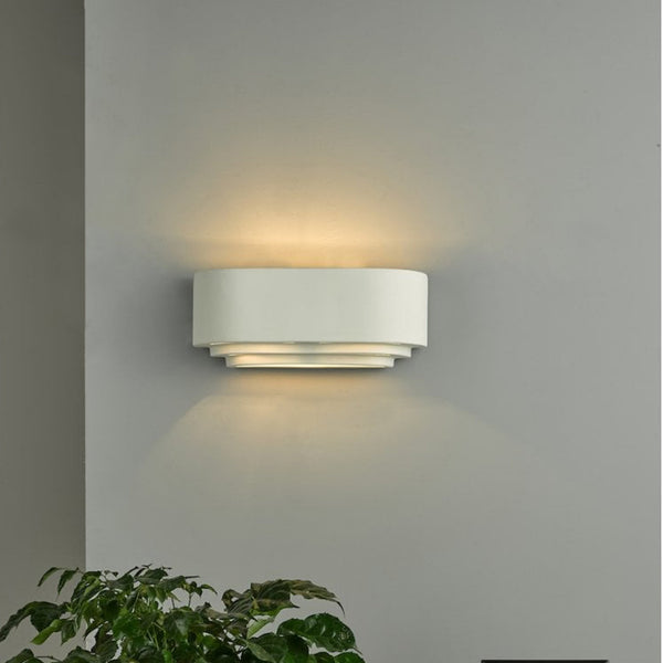 A white oval wall light with tiered base in use, illuminating a wall and plant.