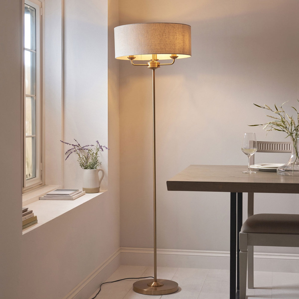 A brass floor lamp with a cream shade illuminates a dining area with a window and a table.