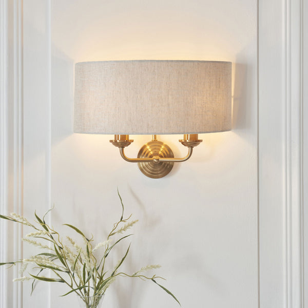 An antique brass wall light with a natural linen drum shade illuminates a white wall.