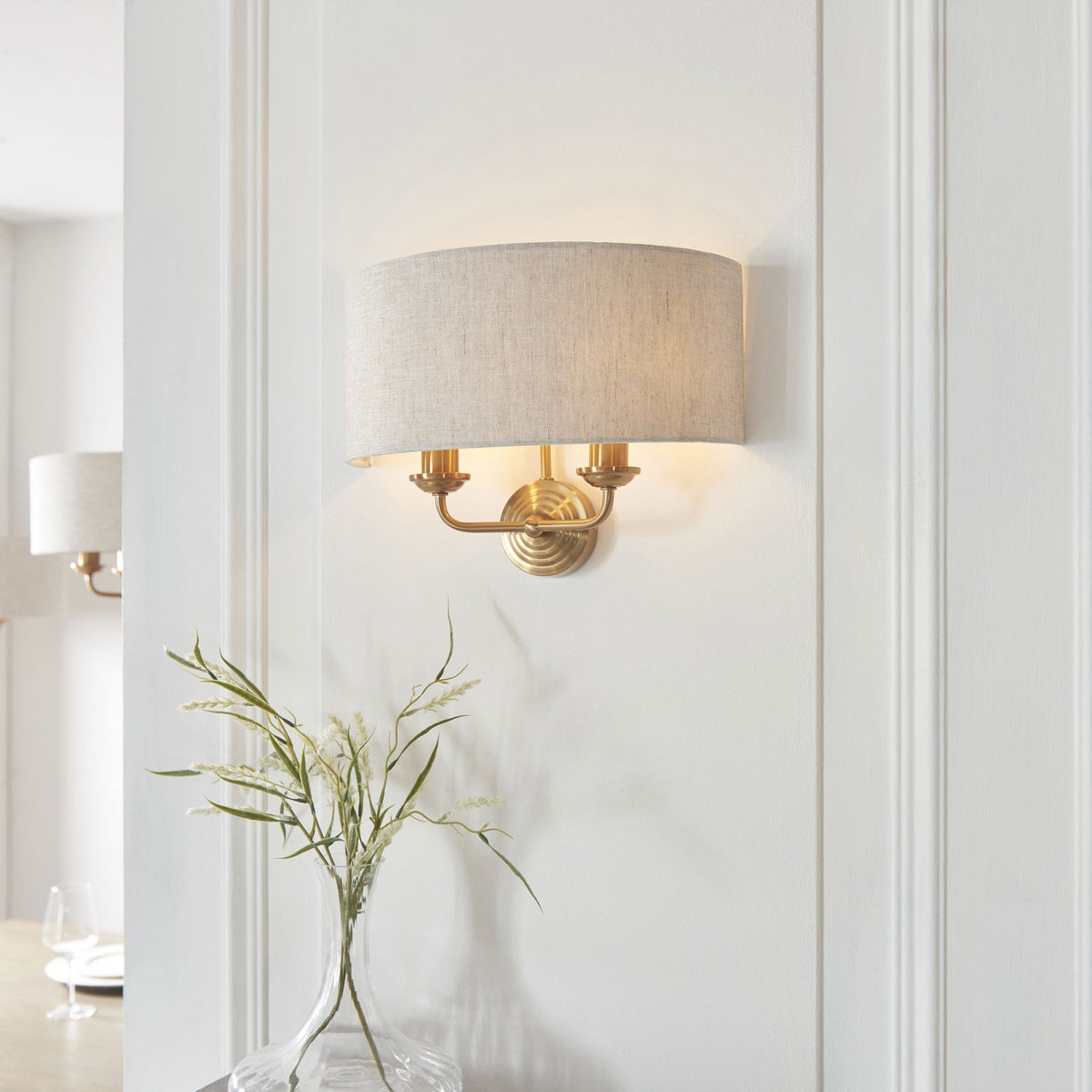 An antique brass wall light with a natural linen drum shade on a white wall in a living room.
