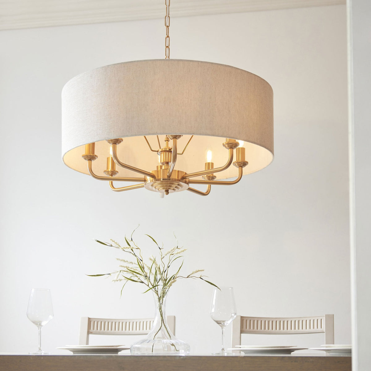 A gold chandelier with a light fabric drum shade hangs above a dining table with a vase of greenery.