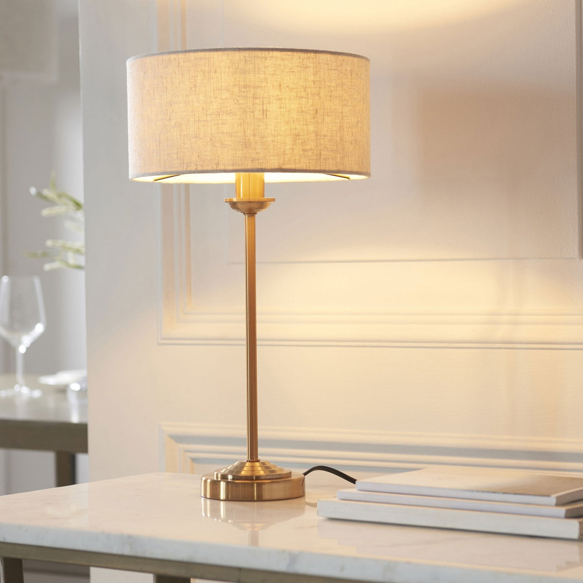 A brass table lamp with a linen shade illuminates a white marble table with books.