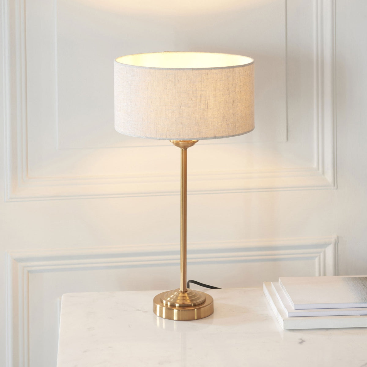 A brass table lamp with a linen shade sits on a white marble table next to a stack of books.