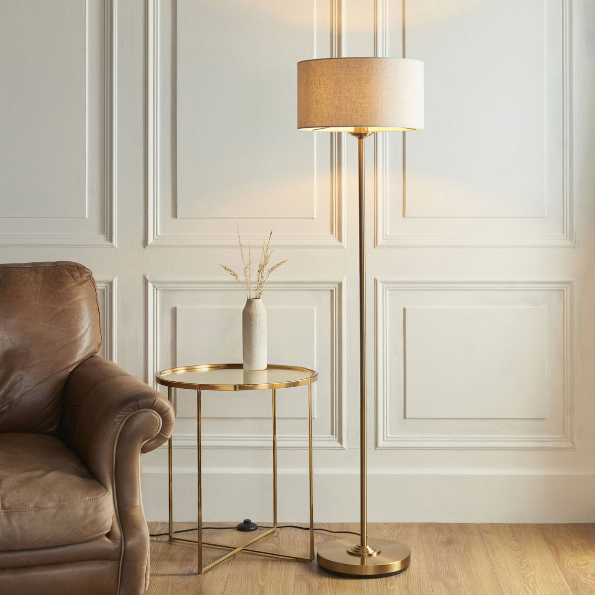 A brass floor lamp with a light linen shade stands next to a brown leather armchair and a round gold side table.