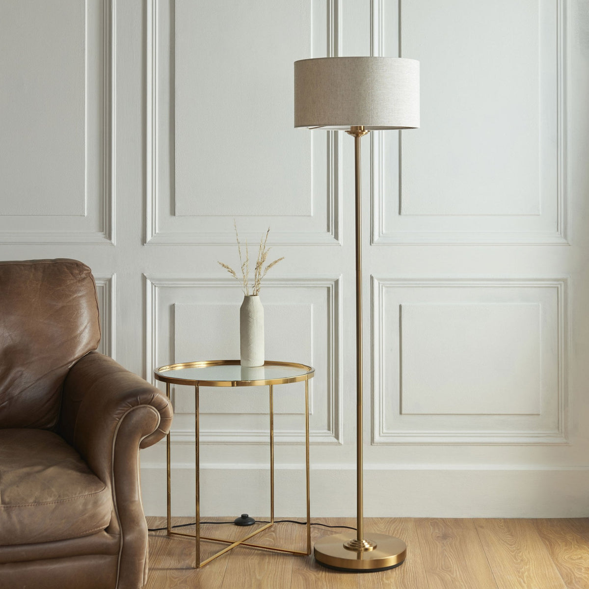 A brass floor lamp with a light linen shade illuminates a cozy living room scene with a leather armchair.