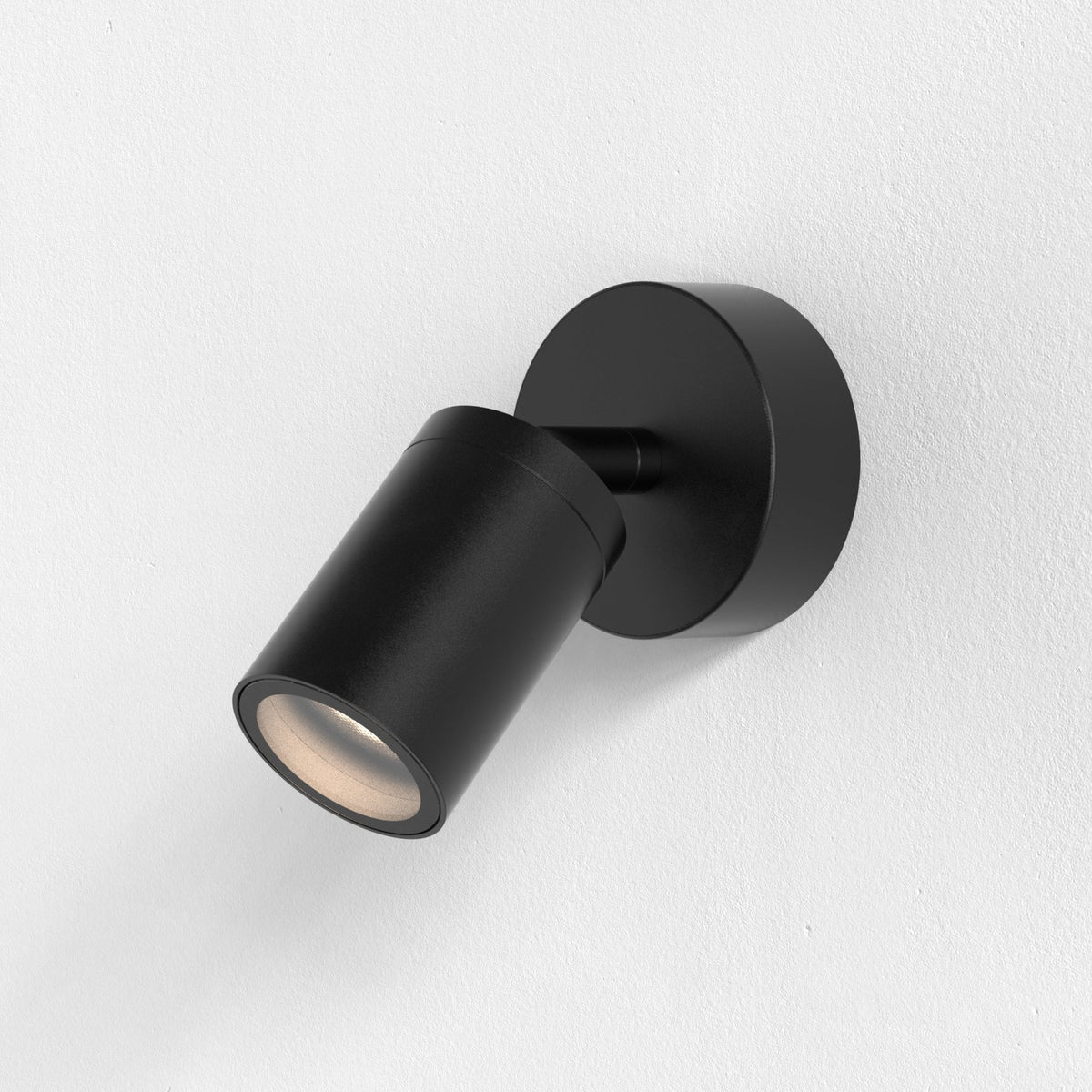 Black cylindrical wall light with a circular base, angled downwards on a white textured wall