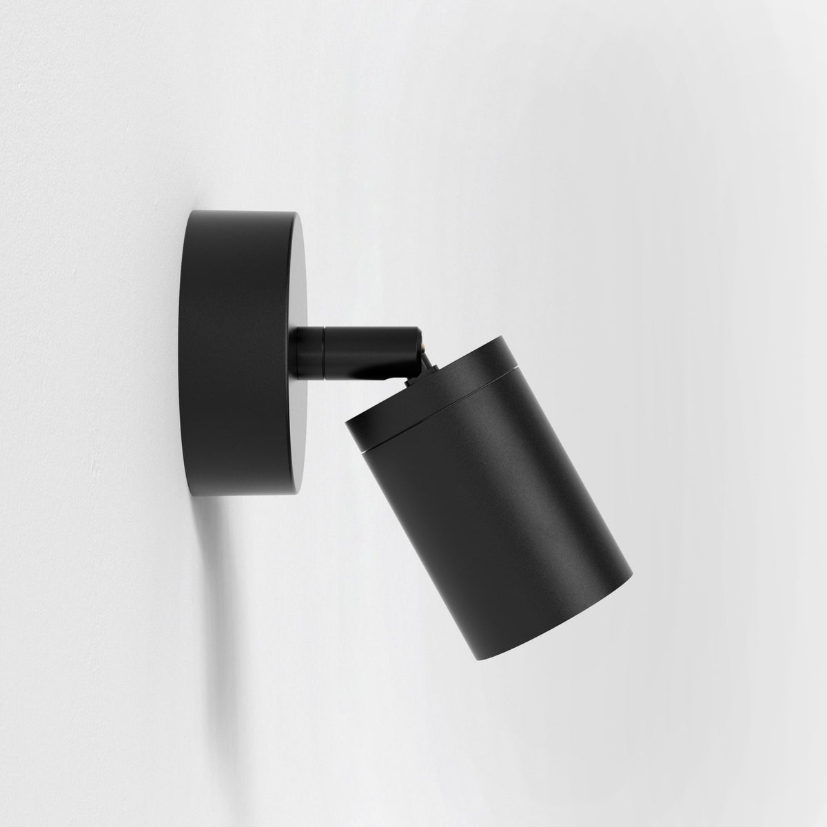 Black cylindrical wall-mounted spotlight with an adjustable head, angled downwards against a white wall