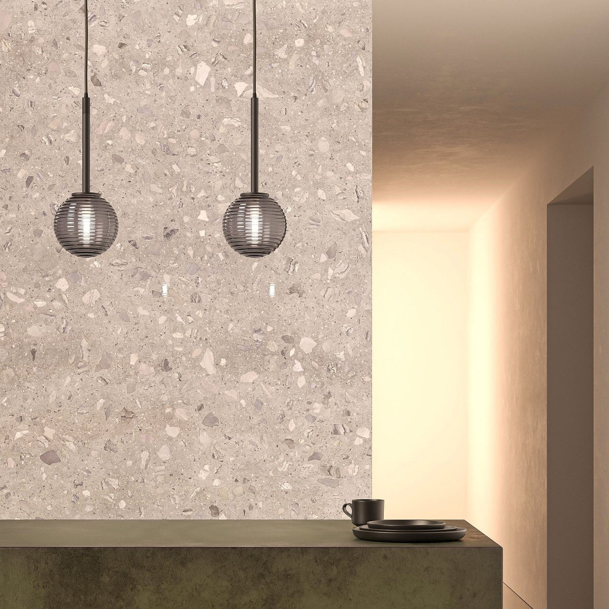 Two pendant lights hang over a dark green counter with black plates and a cup, against a terrazzo wall