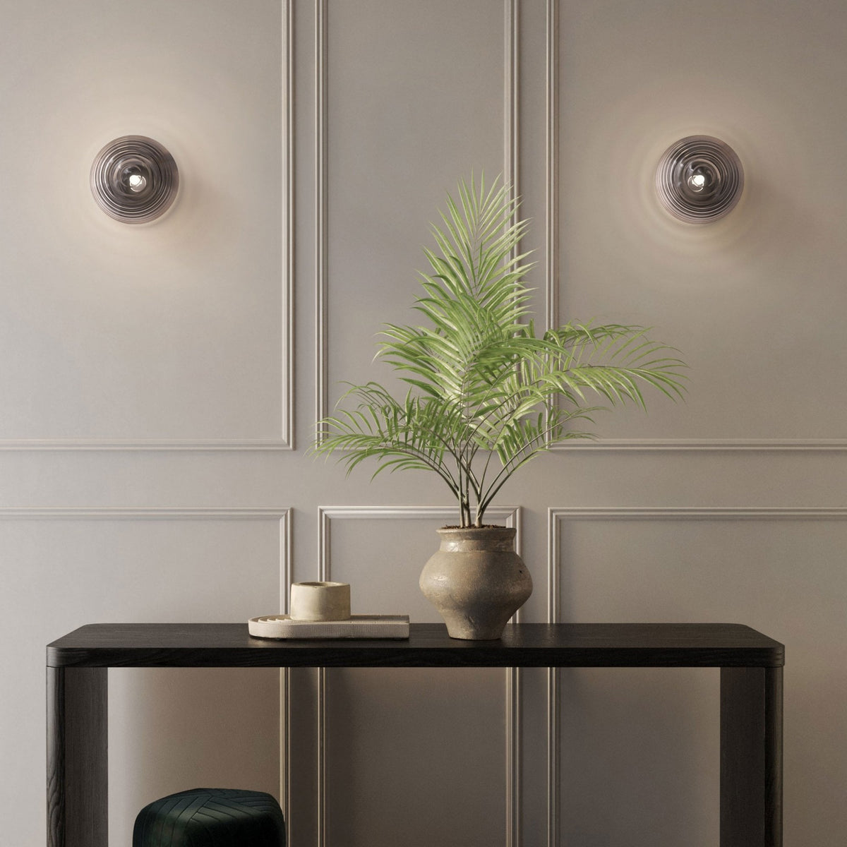 Two grey wall lights illuminate a room with a plant on a dark console table.