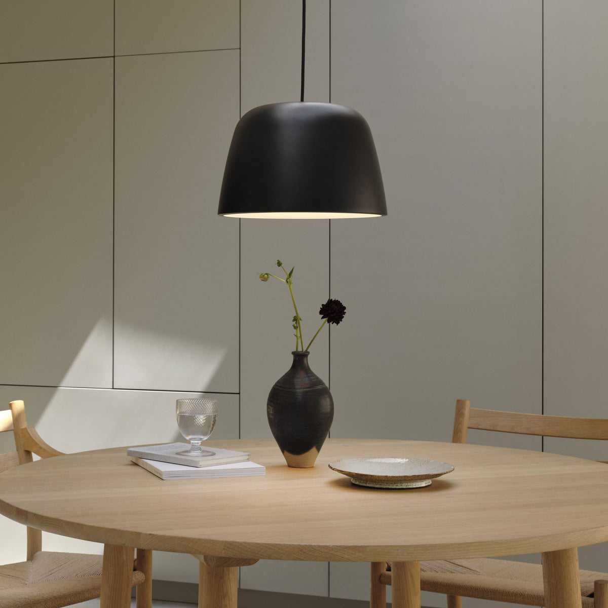 Black pendant light hanging over a light wood round dining table with a vase, glass, and plate