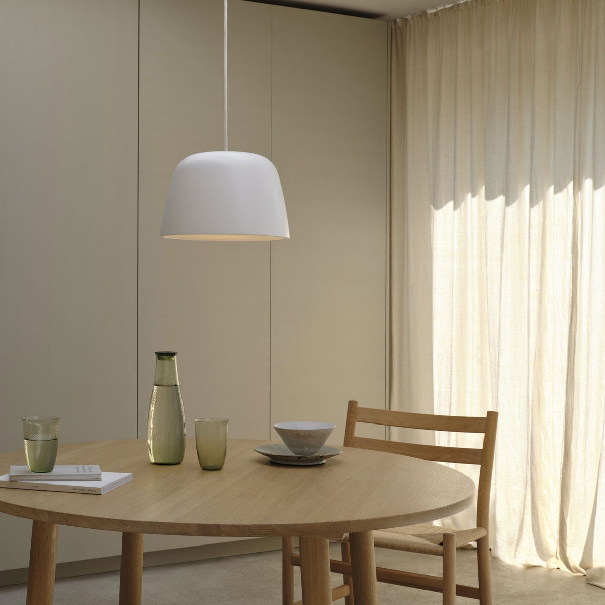 White pendant light above a round wooden dining table with a glass carafe, two glasses, and a bowl