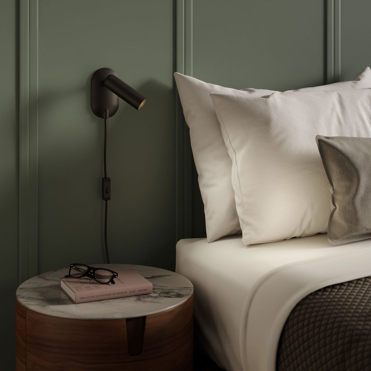 A bedroom with a green wall, a bed with white pillows and a brown quilted blanket, and a side table with a book and glasses