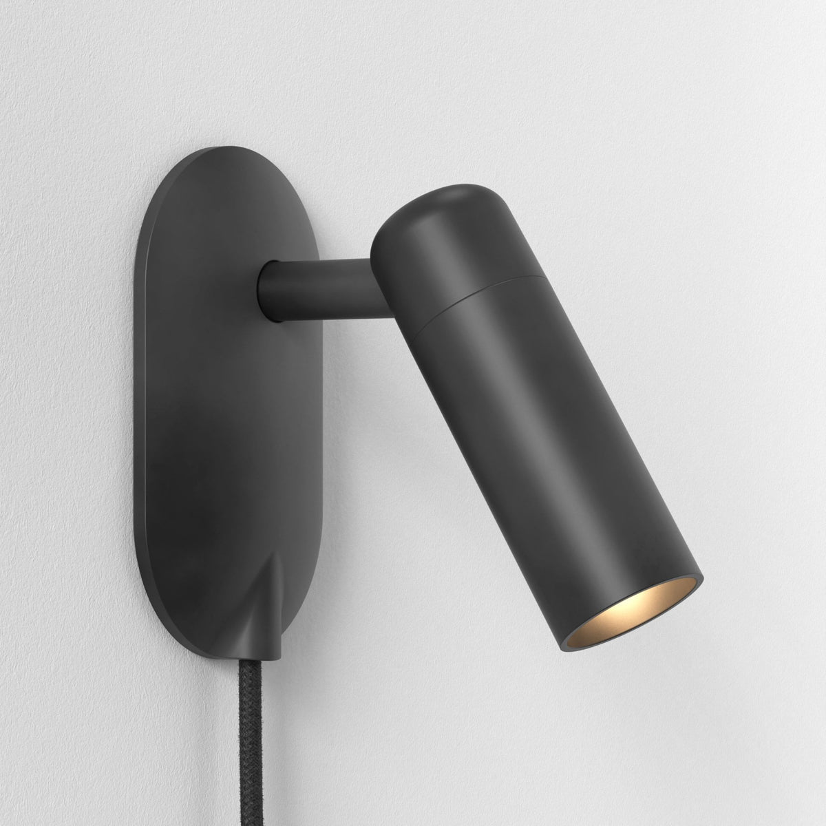 Black wall-mounted spotlight with a cylindrical head angled downwards, emitting a warm glow