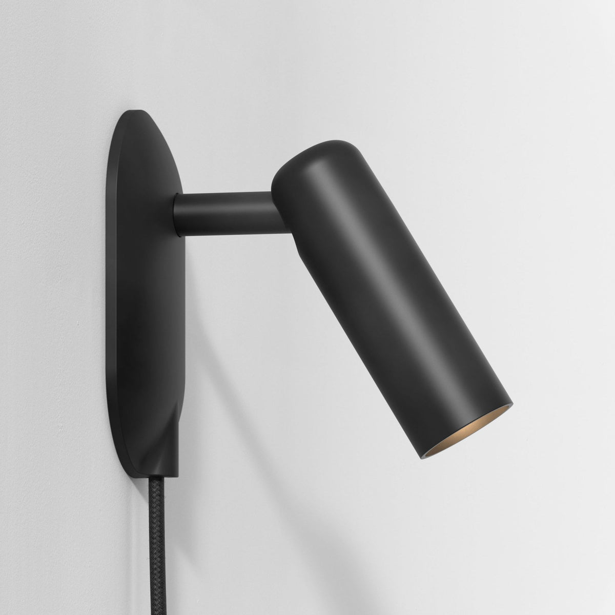 Black wall-mounted light fixture with a cylindrical head angled downwards, revealing a warm-toned interior