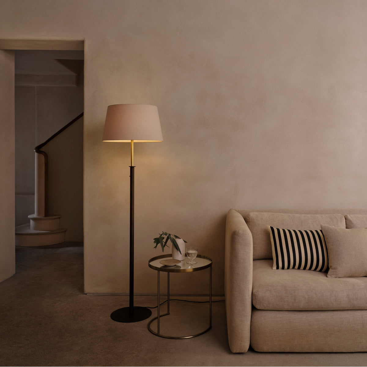 Floor lamp with a beige shade next to a sofa with striped cushions and a side table with a plant