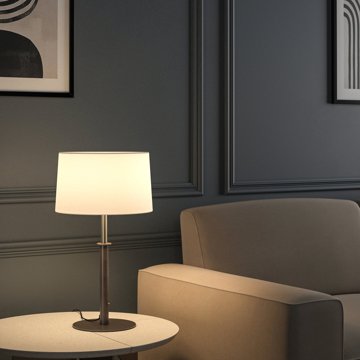 Table lamp with a white shade and dark base on a light round table next to a beige armchair
