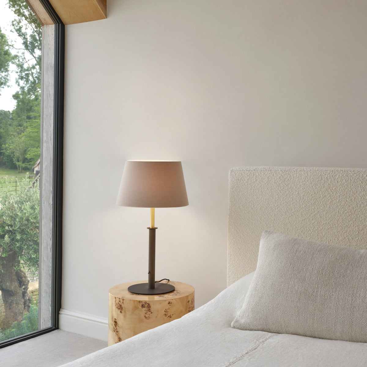 Bedroom with a large window, a bed with a textured headboard, and a table lamp on a wooden stump side table