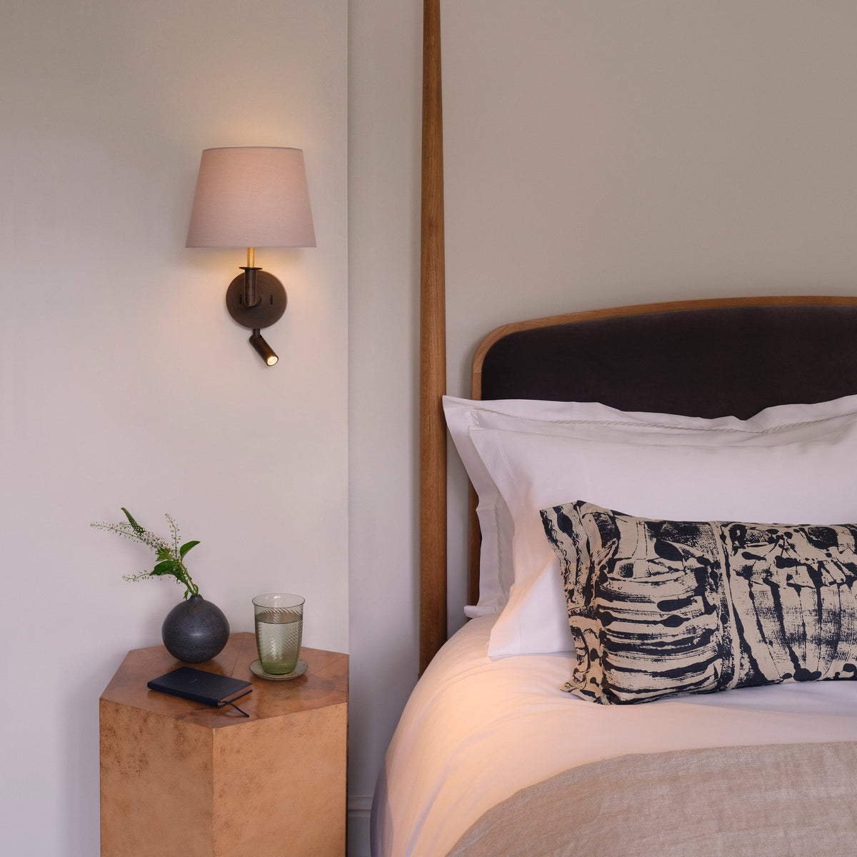 Bedroom with a wooden bed frame, white bedding, and a patterned pillow. A bedside table holds a vase and glass