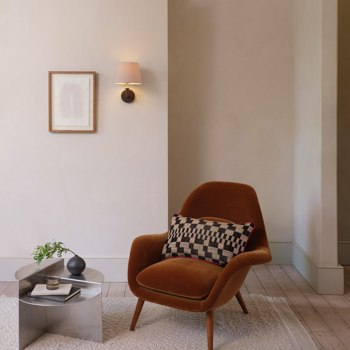 A brown velvet armchair with a patterned cushion next to a silver side table with a plant and books