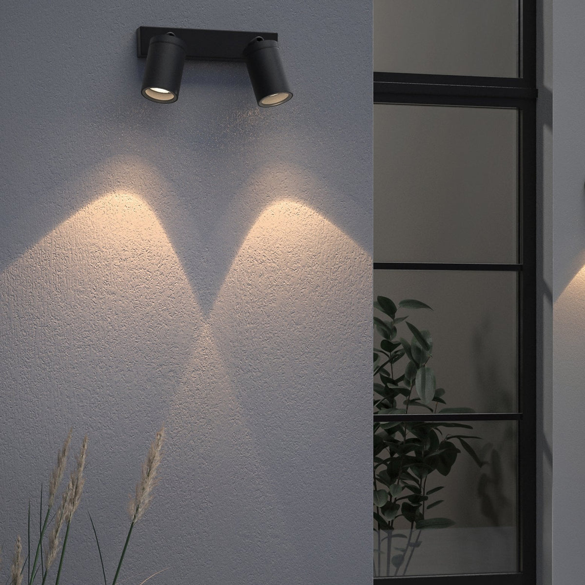 Two black cylindrical outdoor lights mounted on a gray textured wall, illuminating the wall below with warm light