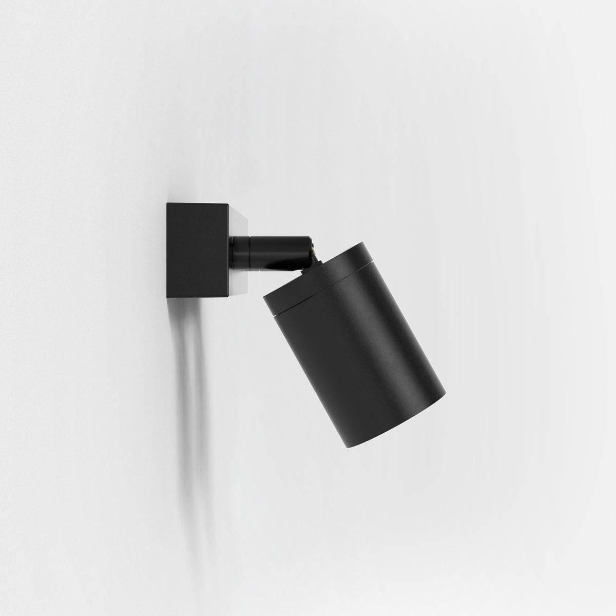 Black wall-mounted spotlight with a cylindrical head angled downwards, casting a shadow on a white wall