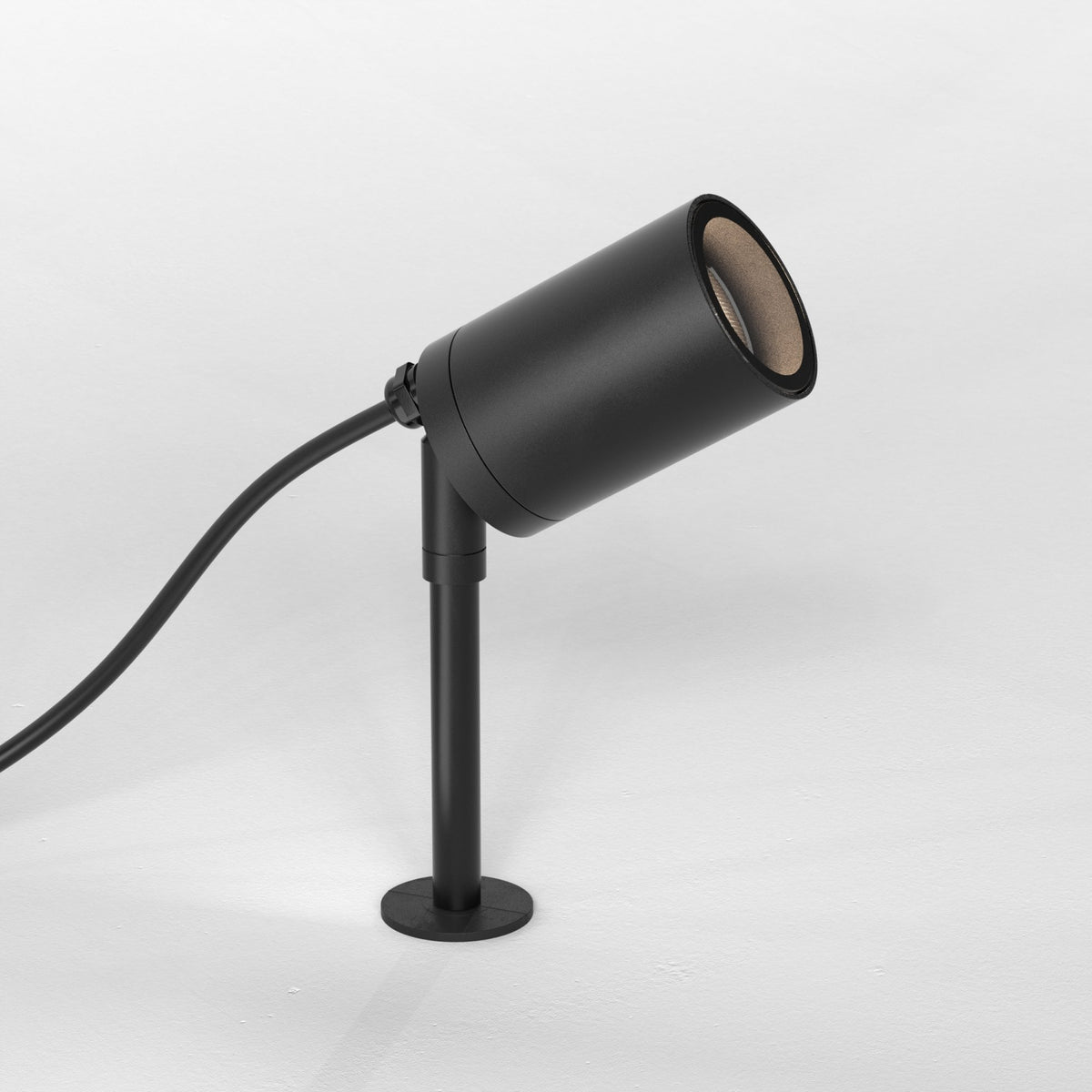 Black cylindrical outdoor spotlight with a round base and a power cord, angled upwards on a white surface