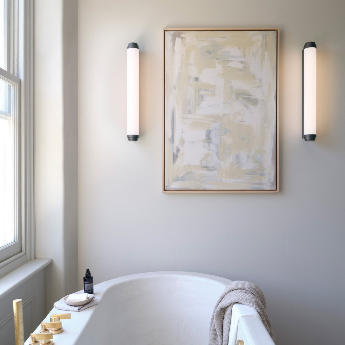 White bathtub with a beige towel, a bottle, and a bar of soap. Two vertical lights flank a painting
