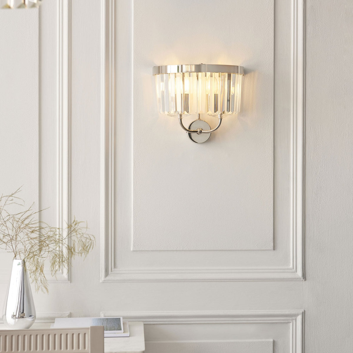 A modern chrome wall sconce with clear glass rods casts a warm glow on a white paneled wall.