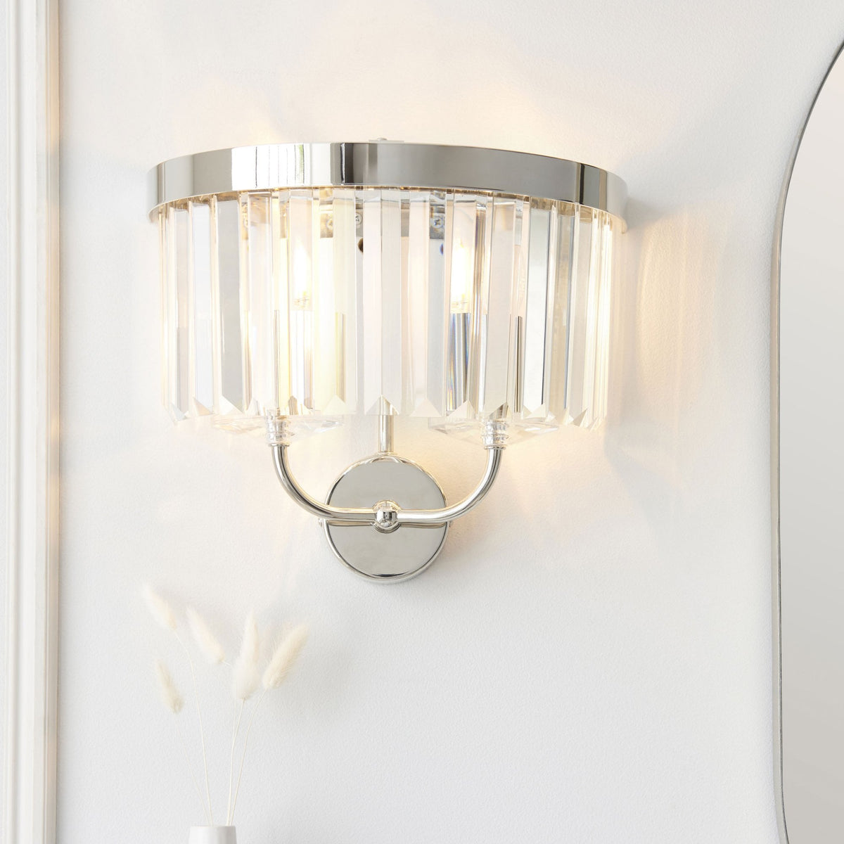 A contemporary chrome wall sconce with vertical glass rods illuminated against a light wall next to a mirror.