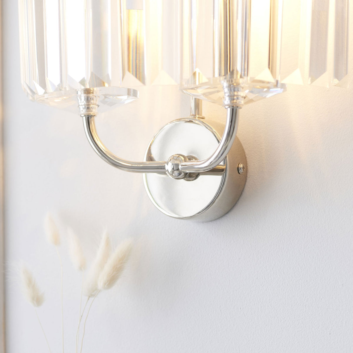 A detailed close-up of the polished chrome base and clear glass rods of a wall light on a white wall.