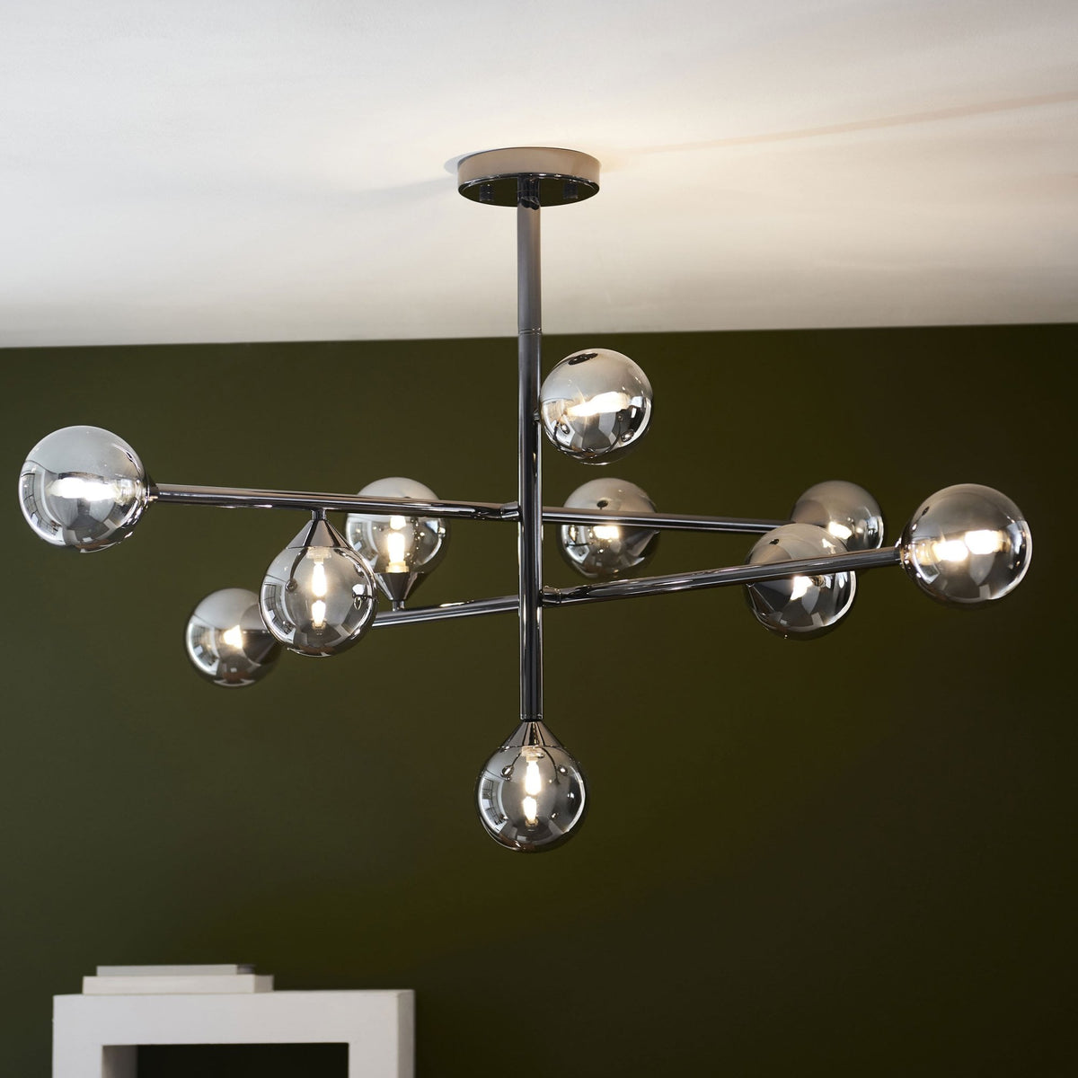 A modern chrome chandelier with multiple arms and spherical smoked glass shades, illuminated against a dark green wall.