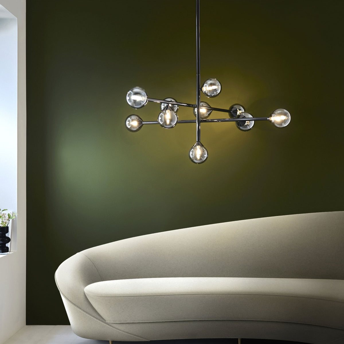 A contemporary chrome chandelier with spherical lights hanging above a curved, light-colored sofa in a room with a dark green wall.