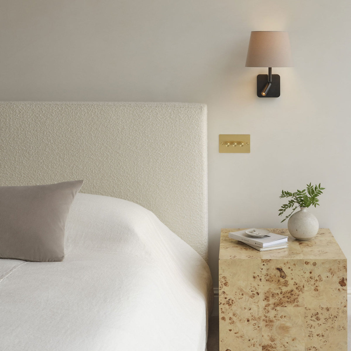 Bedroom with a cream upholstered headboard, a light wood bedside table, and a wall-mounted lamp