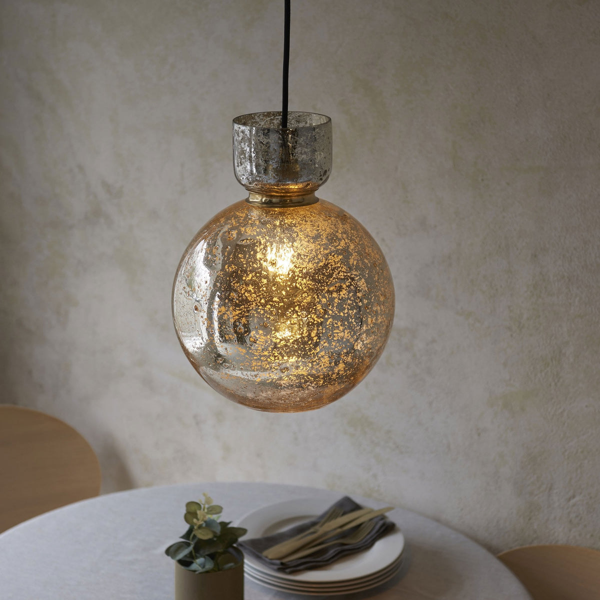 A stylish, round mercury glass pendant light hangs above a minimalist dining table with a plant and plates.