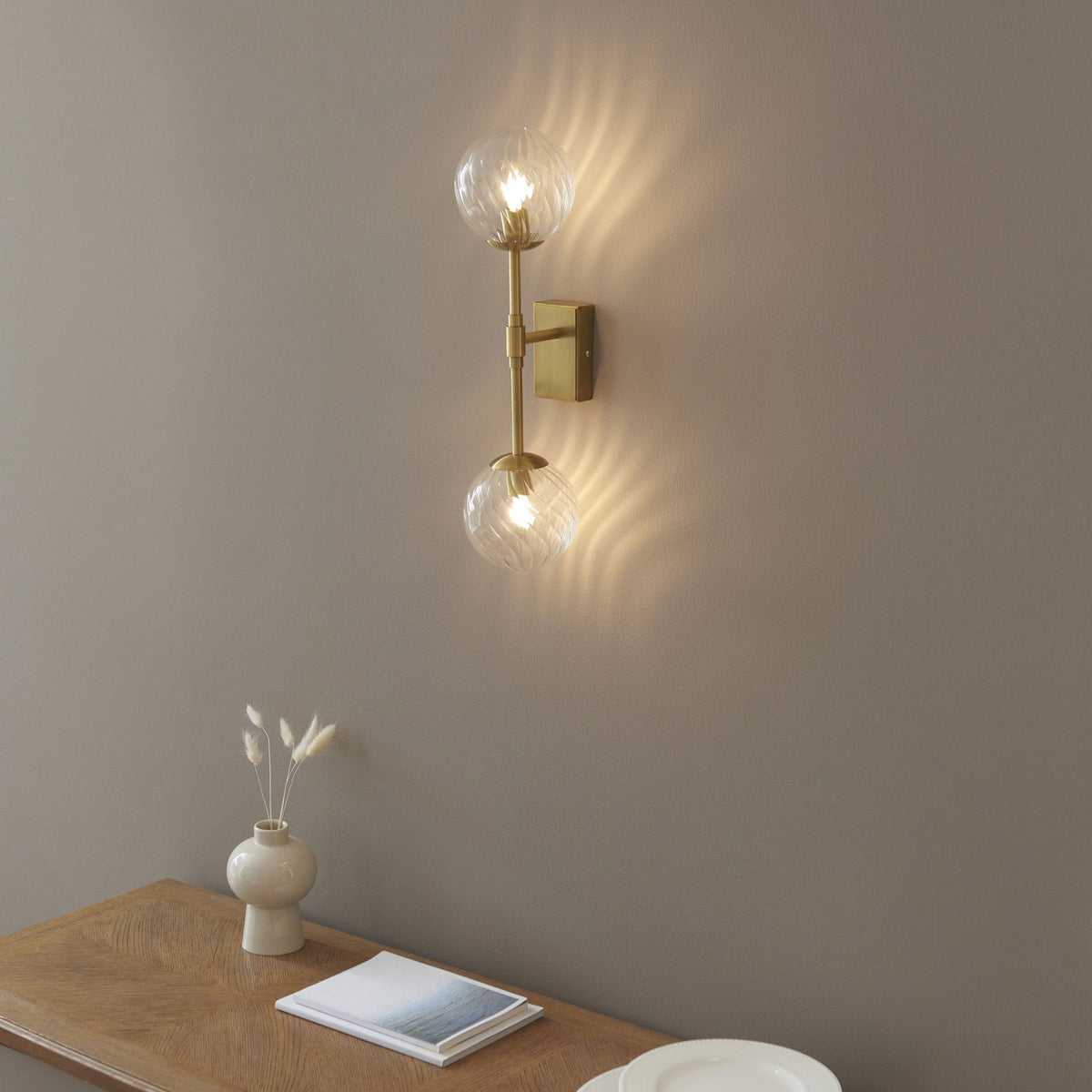 Une applique murale dorée moderne avec deux globes en verre texturé illumine un mur neutre au-dessus d'une table en bois.