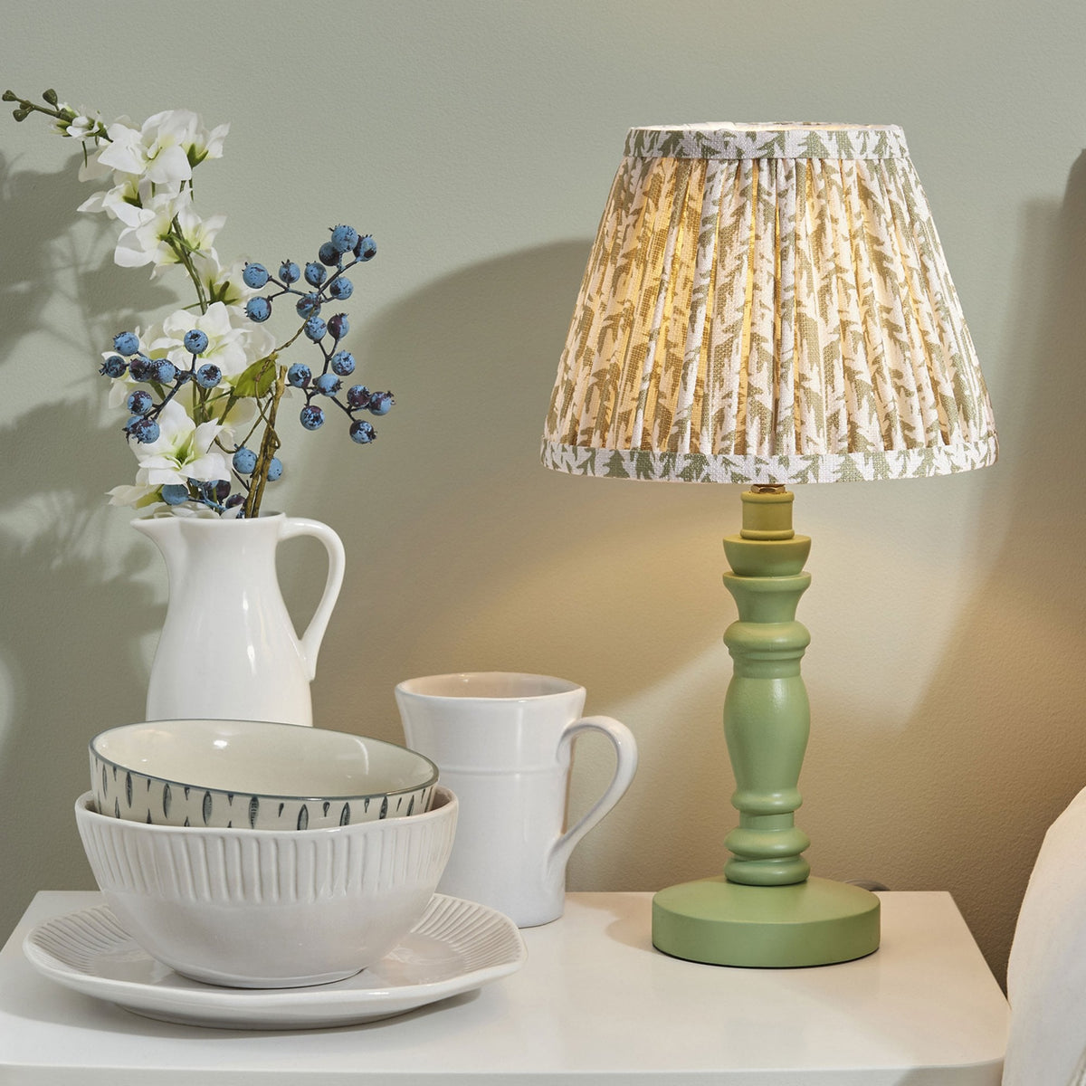 A bedside table lamp with a green wooden base and a patterned shade, next to ceramics and flowers.