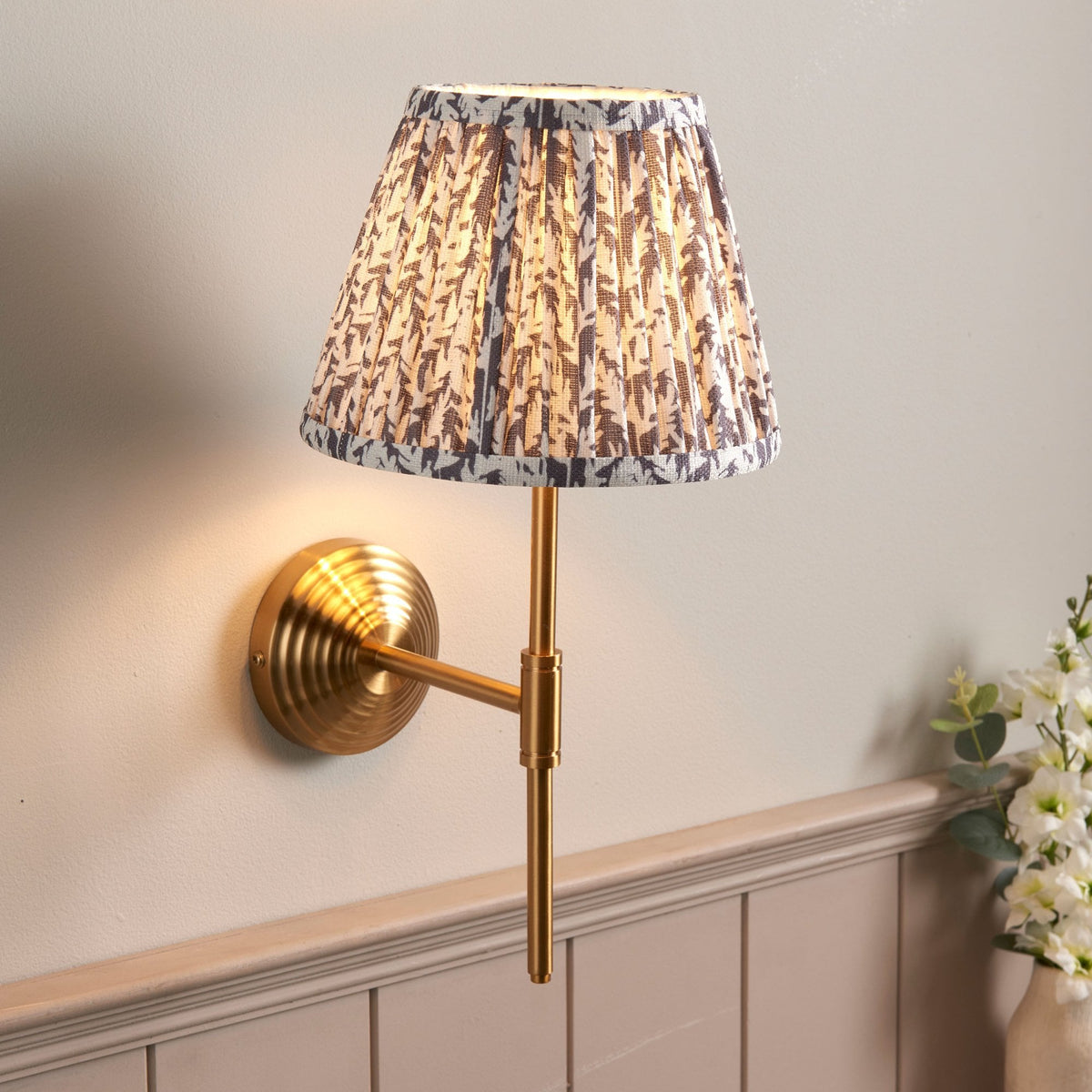 A brass wall light with a grey and white patterned pleated lampshade, illuminated.