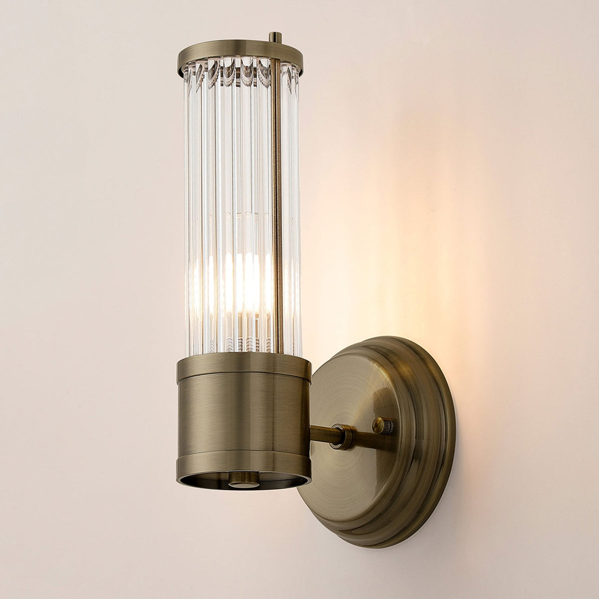 A brass wall light with a fluted glass cylinder shade, illuminated, against a light wall.