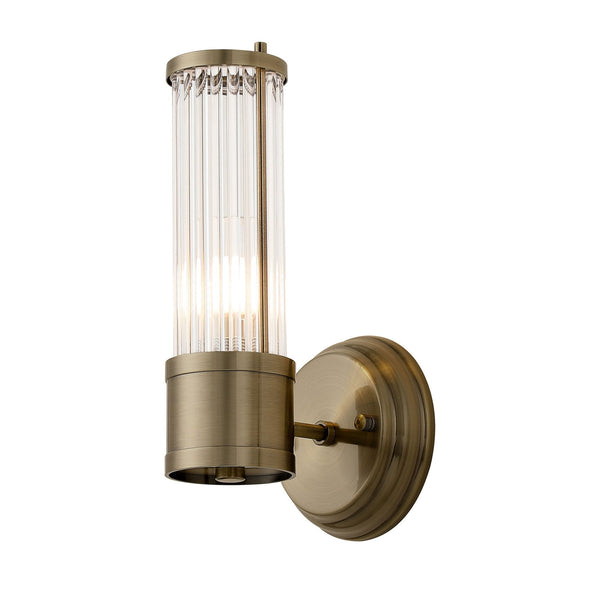 A brass wall light with a fluted glass cylinder shade, illuminated, against a white background.