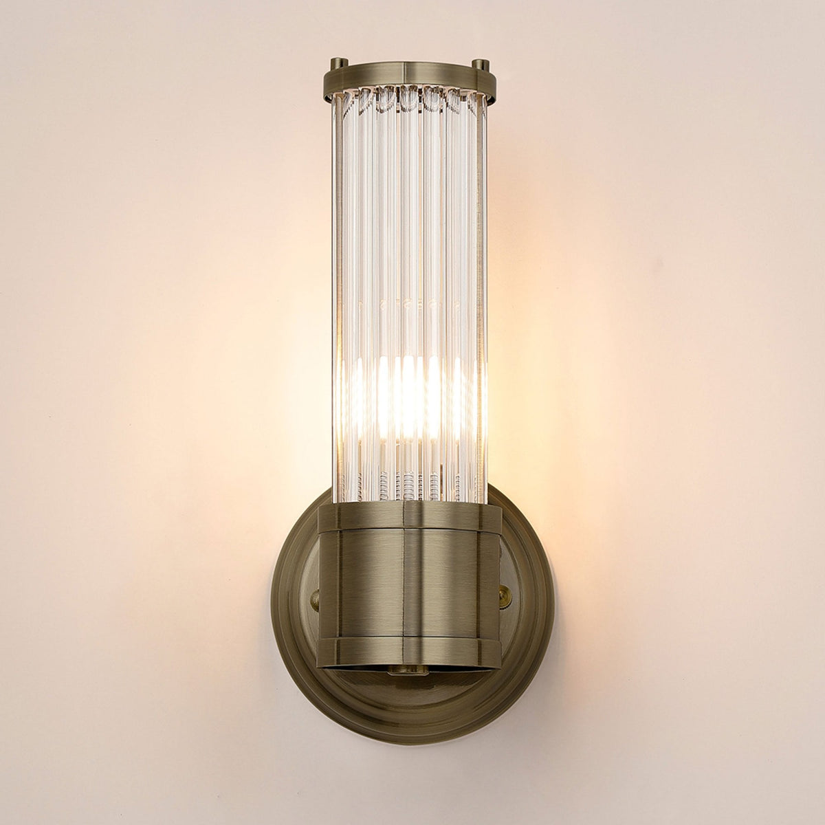 A front view of a brass wall light with a fluted glass cylinder shade, illuminated.