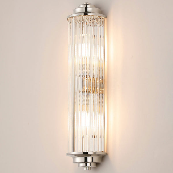 A polished nickel wall light with reeded glass, casting a warm glow on a light wall.