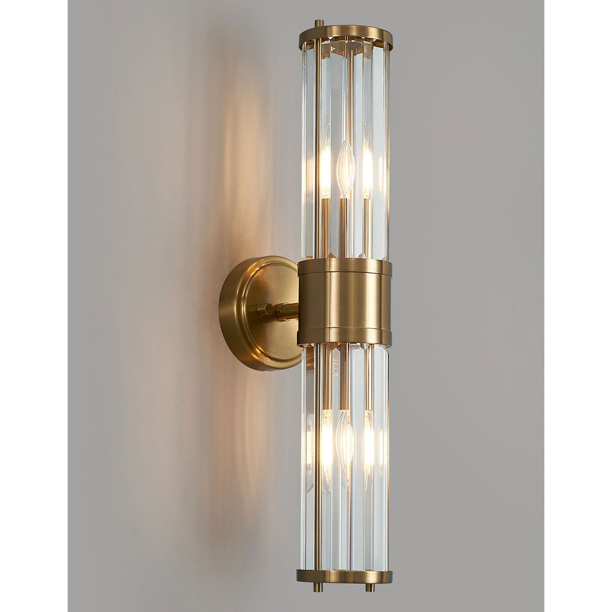A modern brass and fluted glass wall light on a light grey wall, glowing.