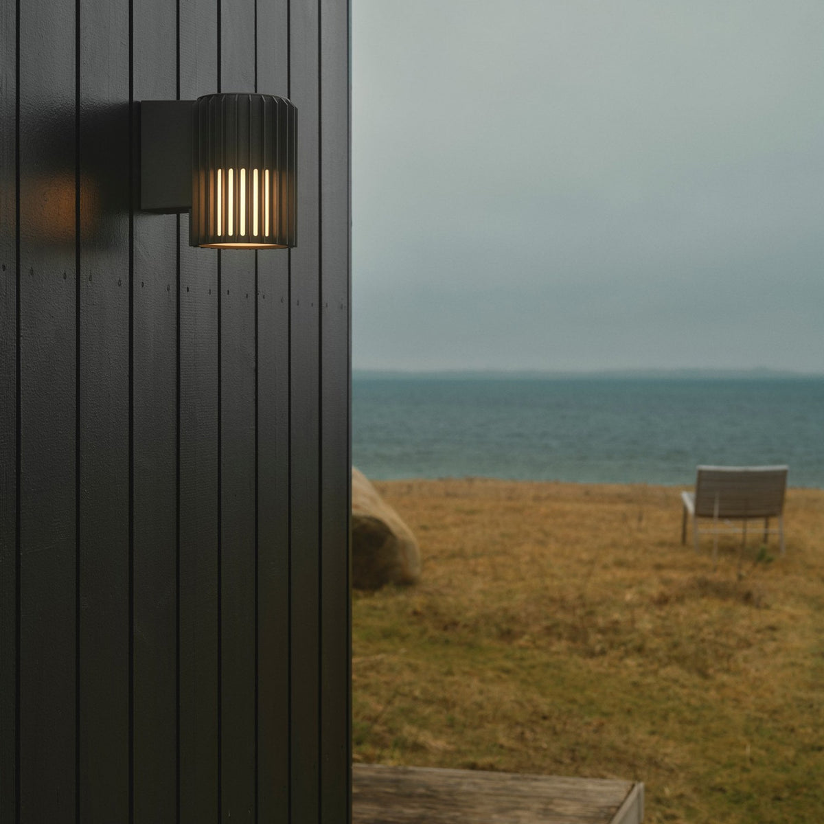 A dark grey outdoor wall light illuminating a modern exterior wall by the sea.