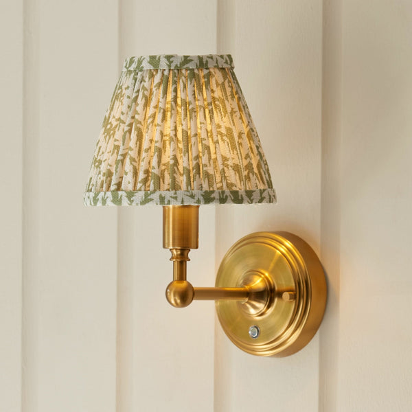A wall sconce with a pleated green and white botanical print shade illuminates a white wall.