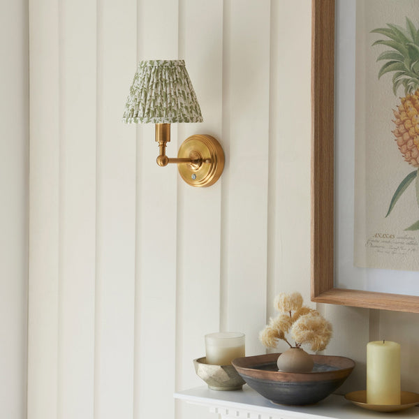 A brass wall sconce with a green patterned shade above a mantelpiece with decor.