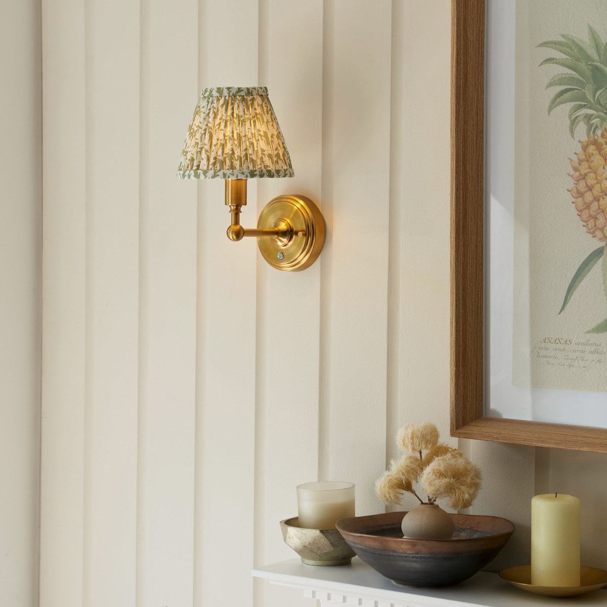 A brass wall sconce with a green patterned shade on a white panelled wall above a mantel.