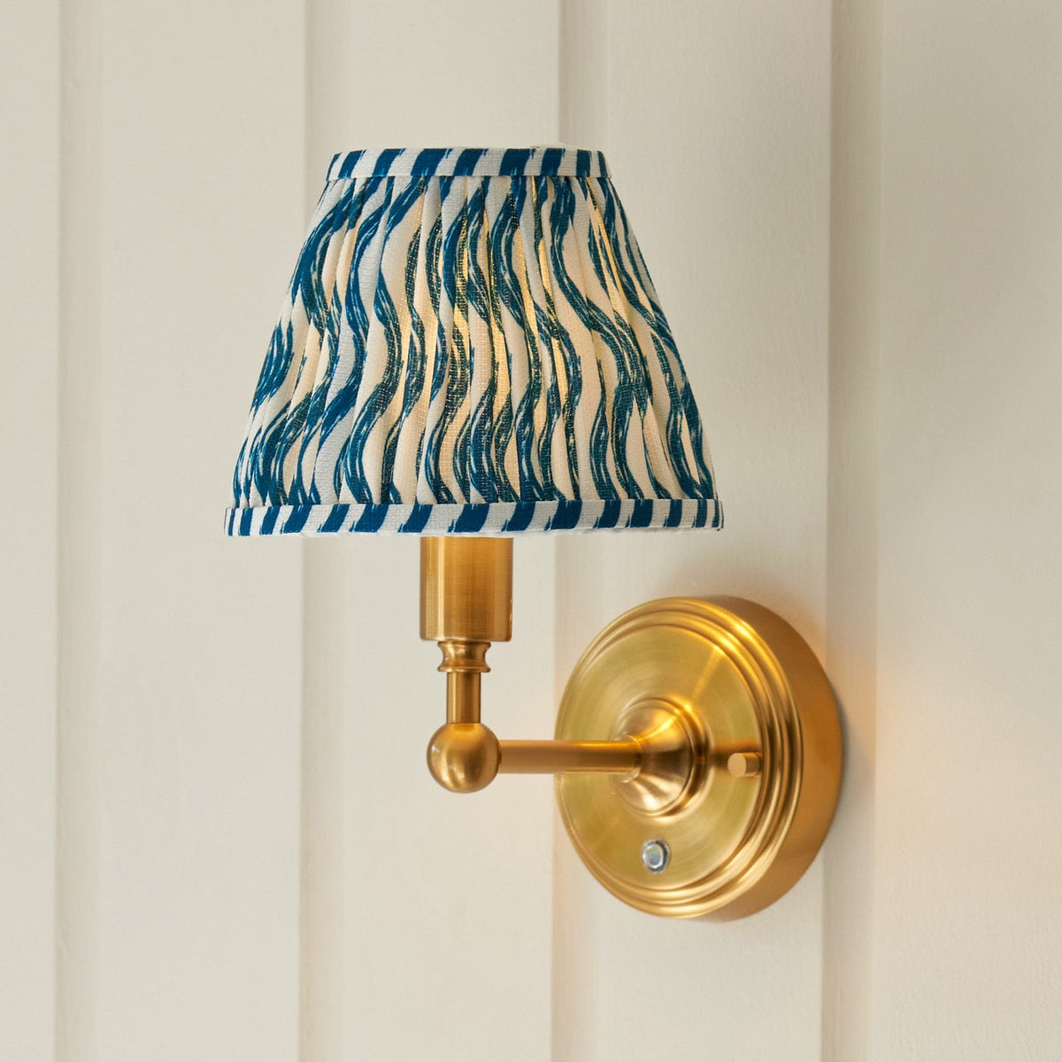 A brass wall light with a blue and white patterned pleated shade on a paneled wall.